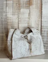 Bolso tipo tote de cuero con acabado craquelado en tonos blanco y beige. Presenta un diseño estructurado con doble asa superior y un cierre frontal mediante cordones de cuero con detalle de borlas.
