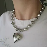 Collar tipo gargantilla plateado con eslabones de cuentas y dijes colgantes: un corazón plateado tridimensional y pequeños cristales incrustados entre las cuentas.