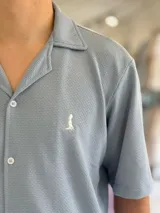 Camisa de manga corta color blanco marfil, confeccionada en tejido texturizado con cuello camisero, cierre frontal de botones y un pequeño bordado oscuro en el pecho.