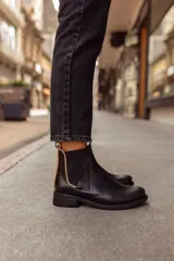 Botas tipo Chelsea de cuero negro, con paneles laterales elásticos y detalle de textura grabada en el talón. Presentan un borde en color beige y tiradores en la parte superior.