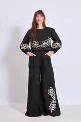 Pantalón negro de corte palazzo con bolsillos laterales y apliques bordados de hojas en color crudo en una pierna.