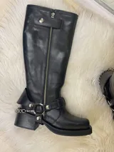 Bota alta tipo biker de cuero negro con cierre metálico lateral que recorre toda la caña. Presenta un diseño versátil con estribo y cadena metálica desmontables en la zona del tobillo, taco cuadrado y plataforma delantera.