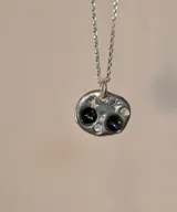 Collar de plata 950 con dije de forma orgánica irregular. El dije presenta un diseño inspirado en el cielo nocturno con dos piedras oscuras circulares, dos pequeñas piedras brillantes y detalles grabados en forma de estrellas.
