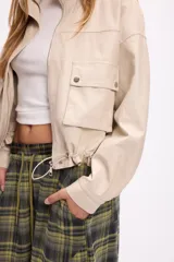 Campera corta de cuero sintético en color beige, con diseño de calce relajado, cierre frontal metálico, bolsillos con solapa en el frente y cordón ajustable en el ruedo.