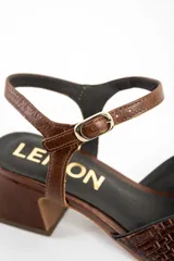 Sandalias de cuero marrón con taco cuadrado medio, tira en el tobillo con hebilla ajustable y diseño trenzado con nudo en la parte delantera.