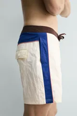Short de baño blanco marfil con cintura marrón y cordón ajustable del mismo color. Presenta franjas laterales en tono azul marino con finos bordes rojizos y bolsillos laterales.