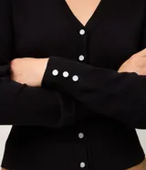 Cardigan de punto blanco con cuello en V, cierre frontal de botones perlados, mangas largas con puños acanalados y detalle de frunces en los hombros. Pertenece a la marca A Collection.