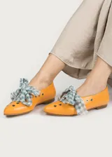 Bailarina de piel natural color naranja con acabado liso, punta redondeada y suela plana. Presenta detalles de ojales metálicos plateados a lo largo del lateral y un lazo desmontable de tela a cuadros vichy en tonos verde agua y blanco sobre el empeine.