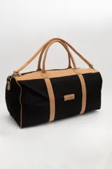 Bolso de viaje tipo duffel de lona negra con detalles en cuero tostado, asas de mano y correa ajustable para el hombro.