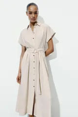 Vestido midi camisero de color beige, con cuello solapa, manga corta y cierre frontal con botones. Incluye un cinturón del mismo tejido con hebilla metálica.