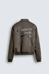 Cazadora estilo varsity de tejido efecto ante en color beige. Presenta cuello solapa, cierre frontal con botones a presión y puños y ruedo con acabado en rib a rayas negras. Incluye parches bordados decorativos en el pecho y la manga, además de bolsillos laterales de doble vivo.