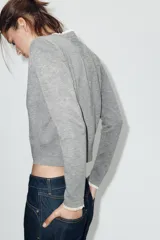 Chaqueta de punto de corte clásico, color gris, con cuello redondo y manga larga. Presenta cierre frontal con botones y detalles de puntilla en color crema en el cuello y los puños.