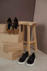Championes urbanos de gamuza color marrón, con detalles en charol negro y bordes dorados. Presentan cordones negros y suela de goma al tono.