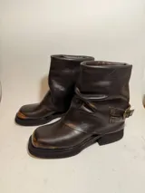 Bota de caña media confeccionada en cuero con efecto desgastado, presenta un diseño holgado en la parte superior y una tira con hebilla metálica ajustable en el tobillo. Cuenta con suela de plataforma y taco cuadrado robusto.