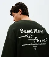 Remera de corte oversized color verde oscuro, con cuello redondo y mangas cortas. Presenta un diseño estampado en la espalda con tipografía en blanco y un pequeño logo gráfico en la parte inferior.