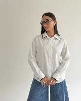 Camisa oversize de corte recto, color blanco, con cuello camisero y puños con detalle de bordado en contraste color negro. Presenta cierre frontal de botones y mangas largas.