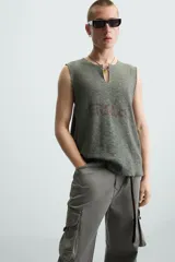 Camiseta sin mangas de punto acanalado, color verde oliva desgastado, con cuello redondo y abertura frontal en forma de 'V'. Presenta un estampado desvanecido en el centro del pecho.