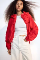 Chaqueta corta de nylon color beige, con cuello subido, capucha y cierre frontal con cremallera. Presenta mangas largas con puños elásticos, bolsillos laterales y detalles de cordones ajustables con lazadas en el pecho y el bajo.