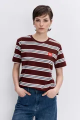 Remera de manga corta con cuello redondo, confeccionada en tejido de punto con diseño de rayas horizontales en tonos marrón, blanco y celeste. Presenta un pequeño parche circular rojo en el pecho.
