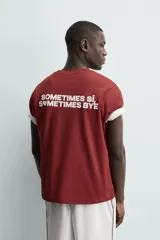 Camiseta blanca de corte relajado (relaxed fit) con cuello redondo y manga corta. Presenta un bolsillo plastrón en el pecho y un estampado de texto en color marrón rojizo en la espalda que dice "SOMETIMES SÍ, SOMETIMES BYE" (reemplazando la 'O' de 'SOMETIMES' por un corazón).