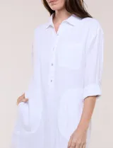 Vestido camisero corto de color blanco con sutiles rayas verticales, de la marca Dash. Presenta cuello con solapa, escote en V con botones parciales en el frente, bolsillo superior en el pecho y bolsillos laterales de gran tamaño. Cuenta con mangas largas de largo regulable mediante presilla y botón, calce holgado y ruedo trasero más largo.