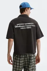 Camisa cargo de manga corta color marrón oscuro, con cuello abierto, botones frontales, bolsillos con solapa en el pecho y el texto 'SERVICE GARMENTS' bordado sobre el bolsillo izquierdo.
