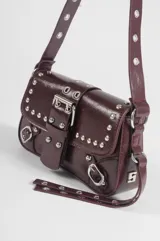 Cartera de hombro de cuero sintético marrón oscuro, con diseño de solapa frontal adornada con tachas metálicas plateadas, hebillas decorativas y correa ajustable.