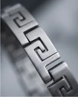 Pulsera rígida de acero inoxidable con diseño de guarda griega en los extremos y placa central lisa apta para grabado.