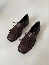 Mocasín de cuero marrón con textura granulada, diseño de horma cuadrada, detalle de flecos y lazo decorativo en el empeine, montado sobre una suela y taco bajo de color negro.