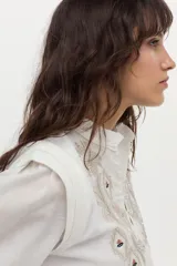 Blusa blanca de tela liviana con bordado detallado en el frente, cuello alto con volado y escote en V con botones. Tiene mangas largas abullonadas con puños amplios y detalles estructurados en los hombros.
