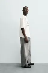 Camiseta blanca de algodón con corte boxy fit y cuello redondo. Presenta un estampado frontal con texto manuscrito en negro y marrón.