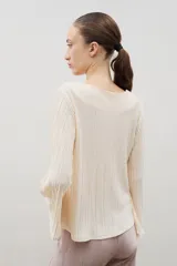 Blusa plisada de color crema con escote en V y mangas largas ligeramente acampanadas. Presenta una textura plisada en toda la prenda que aporta caída y movimiento.