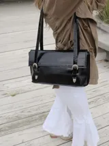 Cartera de hombro color negro con acabado charolado, diseño rectangular con solapa y dos correas fijas. Presenta detalles de hebillas metálicas plateadas en el frente.