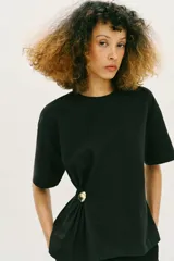 Camiseta de algodón compacto color negro, con cuello redondo, manga corta y detalle de aplique metálico en el frente que genera un ligero fruncido lateral.