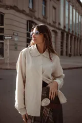 Camisa de lino color beige, con cuello redondo con volados y cierre frontal de botones. Presenta mangas largas con puños abotonados y un corte recto y holgado.