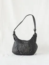 Bolso bandolera de cuero vacuno lavado color negro, con tachuelas plateadas y doradas en el cuerpo. Cuenta con cierre de cremallera, asa de hombro extraíble y bandolera regulable y desmontable.