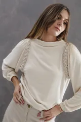 Buzo de lanilla color arena con cuello alto, mangas abullonadas y puños elásticos. Presenta un detalle de cinta tejida trenzada aplicada alrededor de la sisa.