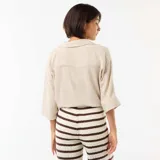 Camisa corta oversize color beige, con mangas largas, cuello camisero, cierre frontal de botones y detalle de nudo en el frente.