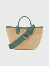 Cartera tipo bandolera de tamaño XS, confeccionada en tejido trenzado de color beige con acabados en cuero blanco. Presenta asas de mano, correa larga ajustable y un parche frontal. Se distingue por un colgante decorativo con cuentas de cerámica en tonos tierra y turquesa.