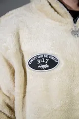 Canguro de peluche color crema, con cuello alto y cierre frontal parcial. Presenta un parche ovalado con diseño gráfico en el pecho y un bolsillo frontal tipo canguro.