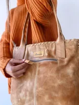 Cartera tipo tote de cuero color camel, con diseño artesanal que presenta costuras decorativas visibles en los laterales y el frente. Cuenta con doble asa de hombro y cierre superior.