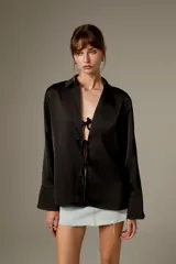 Blusa negra de poliéster, con cuello camisero, mangas largas y cierre frontal mediante tiras para anudar.
