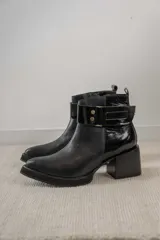 Bota corta de cuero negro con acabado combinado mate y charol. Presenta una tira decorativa con tachas metálicas y hebilla en el tobillo, tacón cuadrado robusto y suela con plataforma.