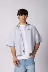 Camisa de manga corta de popelina rayada, con calce boxy y bordado sutil en el pecho. Presenta cierre frontal con botones y diseño abierto.