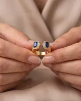 Anillo abierto de bronce con baño de oro, adornado con dos piedras rectangulares de color azul zafiro rodeadas de circonias brillantes y detalles texturizados en los laterales.