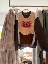 Sweater de diseño artesanal con frente combinado en cuero y gamuza en tonos tierra, decorado con un aplique central geométrico bordado. Presenta mangas y espalda tejidas en punto grueso color marrón oscuro, con detalles de costuras visibles en hilo claro que unen los diferentes materiales.