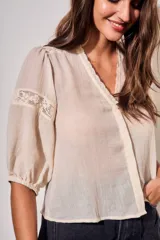Blusa de color arena con cuello en V, mangas tres cuartos abullonadas y detalles de encaje bordado en hombros y mangas. Presenta un diseño ligero y delicado con cierre frontal.