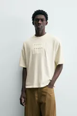 Camiseta de manga corta y cuello redondo, color crema o blanco roto, con un parche texturizado en el pecho que simula barras verticales.