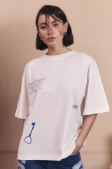 Remerón de corte oversize color off-white, con cuello redondo y mangas cortas. Presenta estampados en color azul que incluyen un dibujo de un edificio, una llave con forma de corazón y la palabra 'renée'.