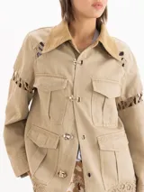 Chaqueta de gabardina liviana color verde militar, con silueta recta, largo por debajo de la cadera y cuello de corderoy. Presenta detalles de encaje en hombros, mangas y ruedo, múltiples bolsillos con solapa y cierre frontal con broches metálicos.